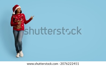 Cheerful pretty african american young woman in red Santa hat posing alone over blue studio background, gesturing, showing copy space for text or advertisement, panorama, full length shot