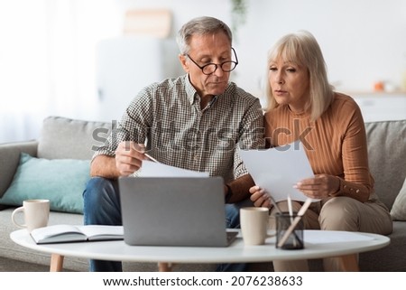 Senior Couple Looking Through Papers Using Laptop Reading Bills And Counting Expenses Together Sitting On Couch At Home. Household And Finances, Retirement Lifestyle Concept Royalty-Free Stock Photo #2076238633