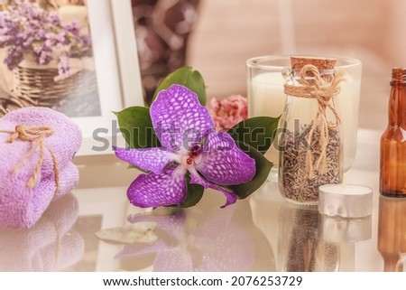 Lavender spa composition. Lilac colored orchid flower , Lilac-colored towel, bottle of lavender herbs, white candle in glass, floral picture in the white frame on glass table. 