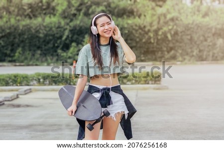 Beautiful sportive hipster happy Asian woman wearing casual shirt and shorts with headphone to listen music while holding skateboard as hobby in free time day on holiday, smiling at outdoor park