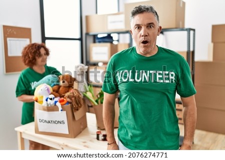 Middle age man wearing volunteer t shirt at donations stand afraid and shocked with surprise expression, fear and excited face. 