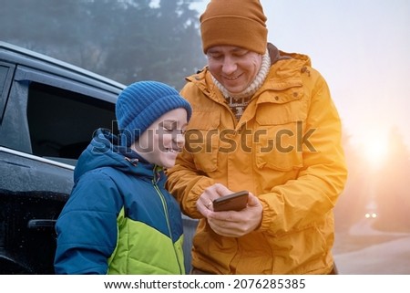Happy father and son standing together near new car on a journey. Family are resting on the side of the road on a road trip. Child takes pictures on smartphone. Happy family travels.
