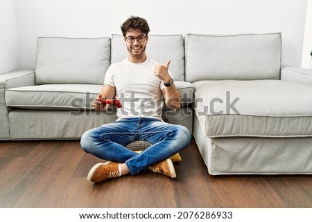 Hispanic man playing video game holding controller by the sofa smiling happy and positive, thumb up doing excellent and approval sign 