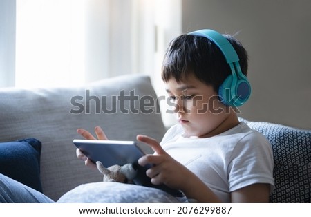 Portrait kid sitting on sofa watching cartoon on tablet, Cute boy sitting next to window playing game online with friends, Cinematic indoor portrait Child wearing headphone listening to music 