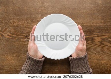 Mature woman hands holding white empty plate on dark brown wooden table background. Closeup. Point of view shot. Meal waiting concept. Royalty-Free Stock Photo #2076302638