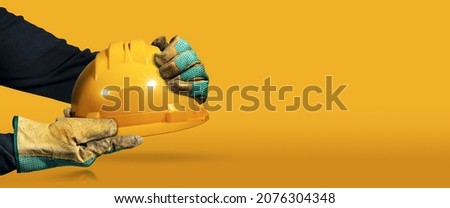 Manual worker with protective work gloves holding a yellow safety helmet on a yellow and orange background with copy space and reflections. Concept of workplace security. Royalty-Free Stock Photo #2076304348