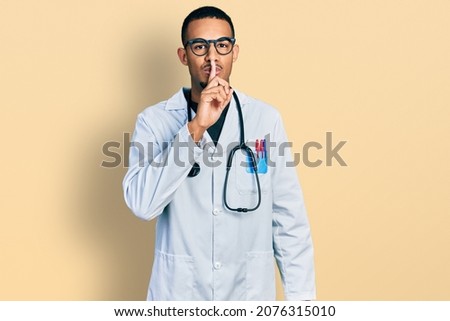 Young african american man wearing doctor uniform and stethoscope asking to be quiet with finger on lips. silence and secret concept.  Royalty-Free Stock Photo #2076315010
