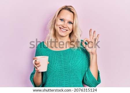 Beautiful caucasian blonde woman drinking a cup of coffee doing ok sign with fingers, smiling friendly gesturing excellent symbol 