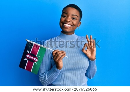 Young african american woman holding kenya flag doing ok sign with fingers, smiling friendly gesturing excellent symbol 