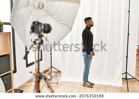Young hispanic man with beard posing as model at photography studio looking to side, relax profile pose with natural face with confident smile. 