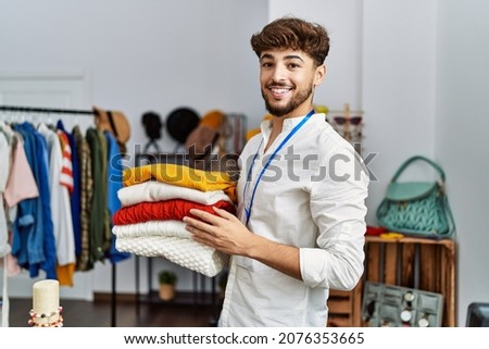 Young arab man shopkeeper holding folded clothes working at clothing store Royalty-Free Stock Photo #2076353665