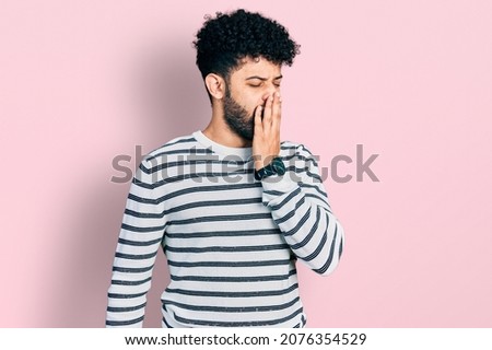 Young arab man with beard wearing casual striped sweater bored yawning tired covering mouth with hand. restless and sleepiness. 