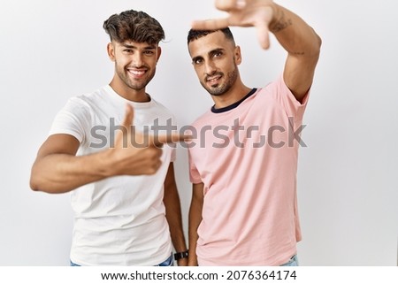 Young gay couple standing over isolated background smiling making frame with hands and fingers with happy face. creativity and photography concept. 