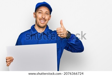 Bald man with beard wearing builder jumpsuit uniform holding empty banner smiling happy and positive, thumb up doing excellent and approval sign 