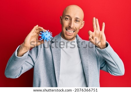 Bald man with beard holding virus toy doing ok sign with fingers, smiling friendly gesturing excellent symbol 