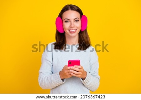 Photo of cheerful positive young woman wear earmuff hold phone repost comment isolated on yellow color background