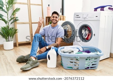 Young hispanic man putting dirty laundry into washing machine showing and pointing up with fingers number three while smiling confident and happy. 