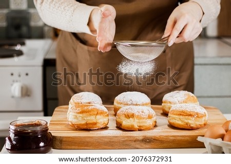 Woman prepares fresh donuts with jam in home kitchen. Cooking traditional Jewish Hanukkah sufganiyot. Hands sprinkle Berliners with powdered sugar. Royalty-Free Stock Photo #2076372931