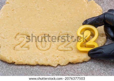 A hand in a black glove cutting out the date 2022 from raw dough for New Year's and Christmas cookies, top view.