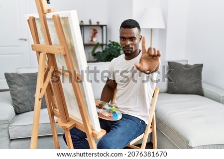 Young african man painting on canvas at home pointing with finger up and angry expression, showing no gesture 