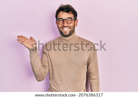 Handsome man with beard wearing turtleneck sweater and glasses smiling cheerful presenting and pointing with palm of hand looking at the camera. 