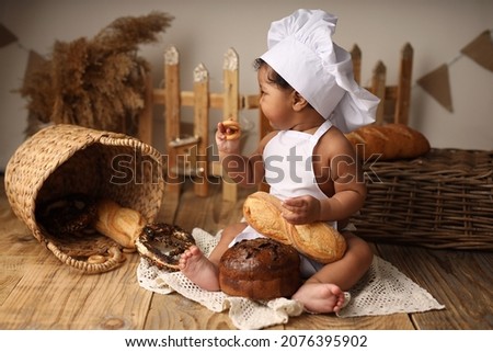 A cute dark-skinned kid with curly hair in a chef's costume eats a bagel and a roll. Wooden natural background. Environmental friendliness. High-quality photography