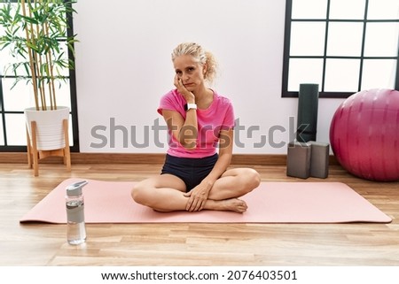 Middle age blonde woman sitting on yoga mat thinking looking tired and bored with depression problems with crossed arms. 