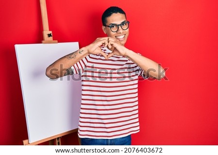 Young african american guy standing by painter easel stand smiling in love doing heart symbol shape with hands. romantic concept. 