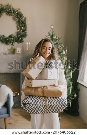 Young beautiful woman holding gift boxes and wrapped presents in a cozy room with Christmas tree on background. Holiday preparations.