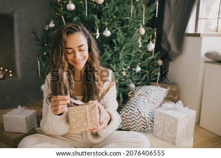 Young beautiful woman holding gift box in a cozy room with Christmas tree and presents on background. Holiday preparations.