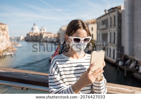 Young woman in medical mask traveling during pandemic in Italy. Concept of social rules of wearing a mask during a pandemic. Woman using smartphone while standing on the bridge in Venice