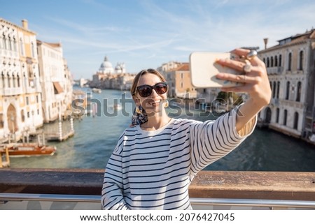 Young woman taking selfie photo on the background of famous Grand Canal in Venice. Idea of spending summer time and happy travel in Italy. Caucasian female in white sunglasses and striped vest