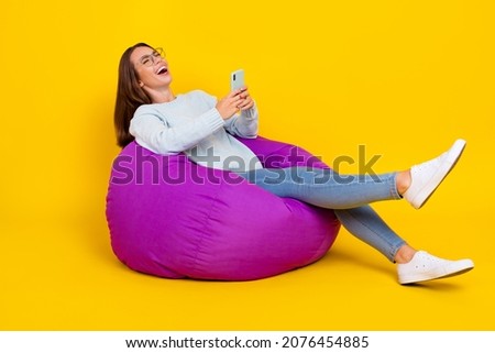 Full length photo of happy cheerful woman enjoy sit chair hold phone laugh joke comment isolated on yellow color background