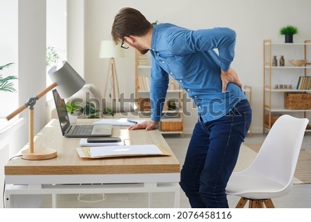 Tired man feels spasm and intense back pain as he stands up after working on computer in sedentary posture for long time. Unhappy stressed young employee has radiculitis or pinched nerve inflammation Royalty-Free Stock Photo #2076458116