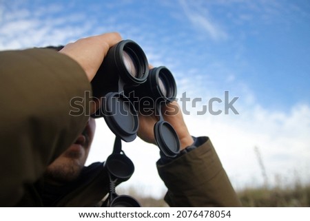 Young man (unshown face) looking into binoculars with blue sky background. Caucasian male observing wildlife outdoors. Nature photographer searching for animals holding binoculars with both hands.  Royalty-Free Stock Photo #2076478054