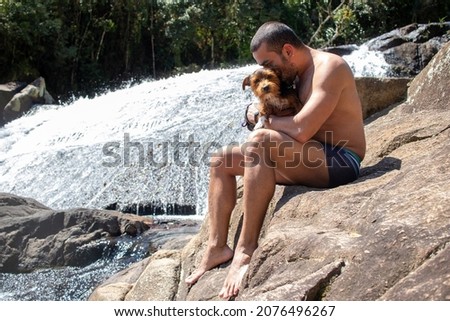 man sitting on stone kissing his little black dog, admiring the waterfall in sunny day.