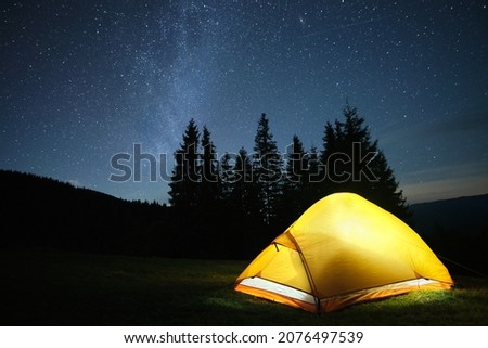 Brightly illuminated camping tent glowing on campsite in dark mountains under night stars covered sky. Active lifestyle and traveling concept.