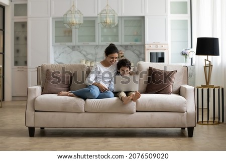 Happy young Indian mother and excited little son playing virtual online videogame on laptop in living room. Mom and kid making video call on computer, resting on couch in pale home interior. Wide shot Royalty-Free Stock Photo #2076509020