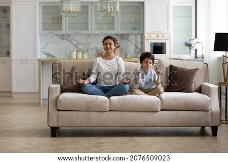 Happy calm Indian mom and little son kid sitting on couch, keeping lotus pose, zen hand gesture, meditating, practicing home yoga, home yoga, mental control, mindfulness, relaxing, exercising Royalty-Free Stock Photo #2076509023