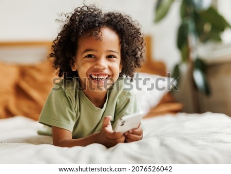 Cute little african american boy with smartphone lying on bed at home, happy child watching cartoons or playing mobile games on phone during leisure time. Gadget addiction among children