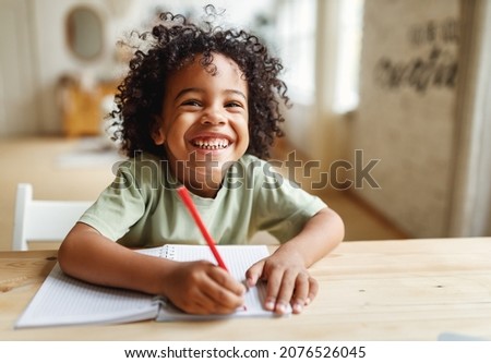 Smiling african american child school boy doing homework while sitting at desk at home, happy mixed race kid practicing handwriting in notebook, learning to write in exercise book Royalty-Free Stock Photo #2076526045
