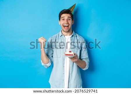 Cheerful man looking happy, celebrating birthday in party hat, holding b-day cake and make fist pump, standing over blue background