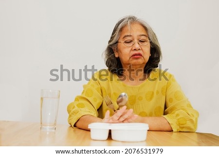 Elderly Asian woman is unhappy and fed up with lunch, a plastic boxed prepackaged meal bought at a convenience store that has no plain taste, sitting on a table with no appetite.
 Royalty-Free Stock Photo #2076531979