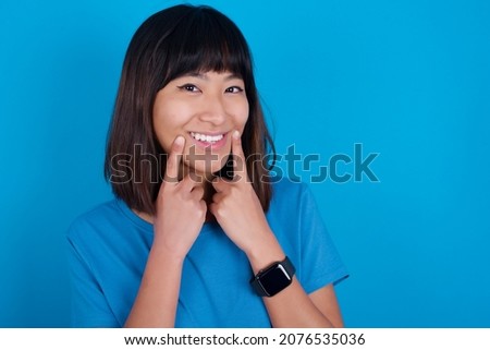Happy young asian woman wearing blue t-shirt against blue background with toothy smile, keeps index fingers near mouth, fingers pointing and forcing cheerful smile