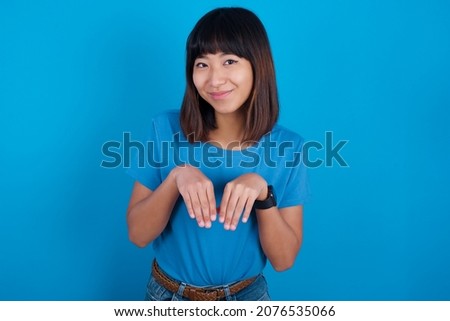 young asian woman wearing blue t-shirt against blue background makes bunny paws and looks with innocent expression plays with her little kid