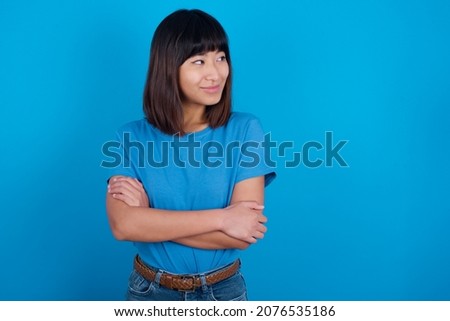 Pleased young asian woman wearing blue t-shirt against blue background keeps hands crossed over chest looks happily aside
