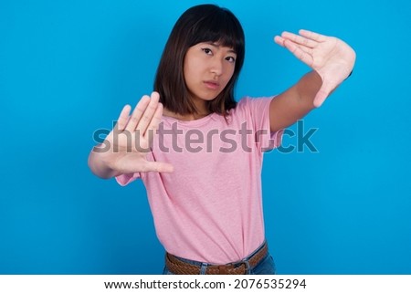 Portrait of smiling young asian woman wearing pink t-shirt against blue background looking at camera and gesturing finger frame. Creativity and photography concept.