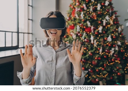 Impressed europian lady using VR googles while spending Christmas at home, playing 3D games in room with beautiful lush Xmas tree on background, happy woman completely immersed in virtual world