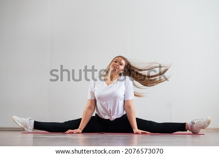 A young flexible fat woman sits in a transverse twine and waves her hair against a white background