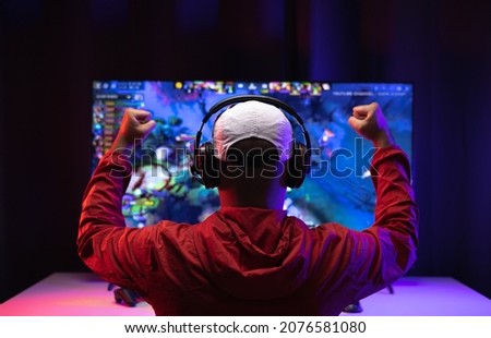 Rear view young Player video computer pc. Young man sitting on chair in game station.Streamer man wearing headphone  playing action game online in the darkroom.